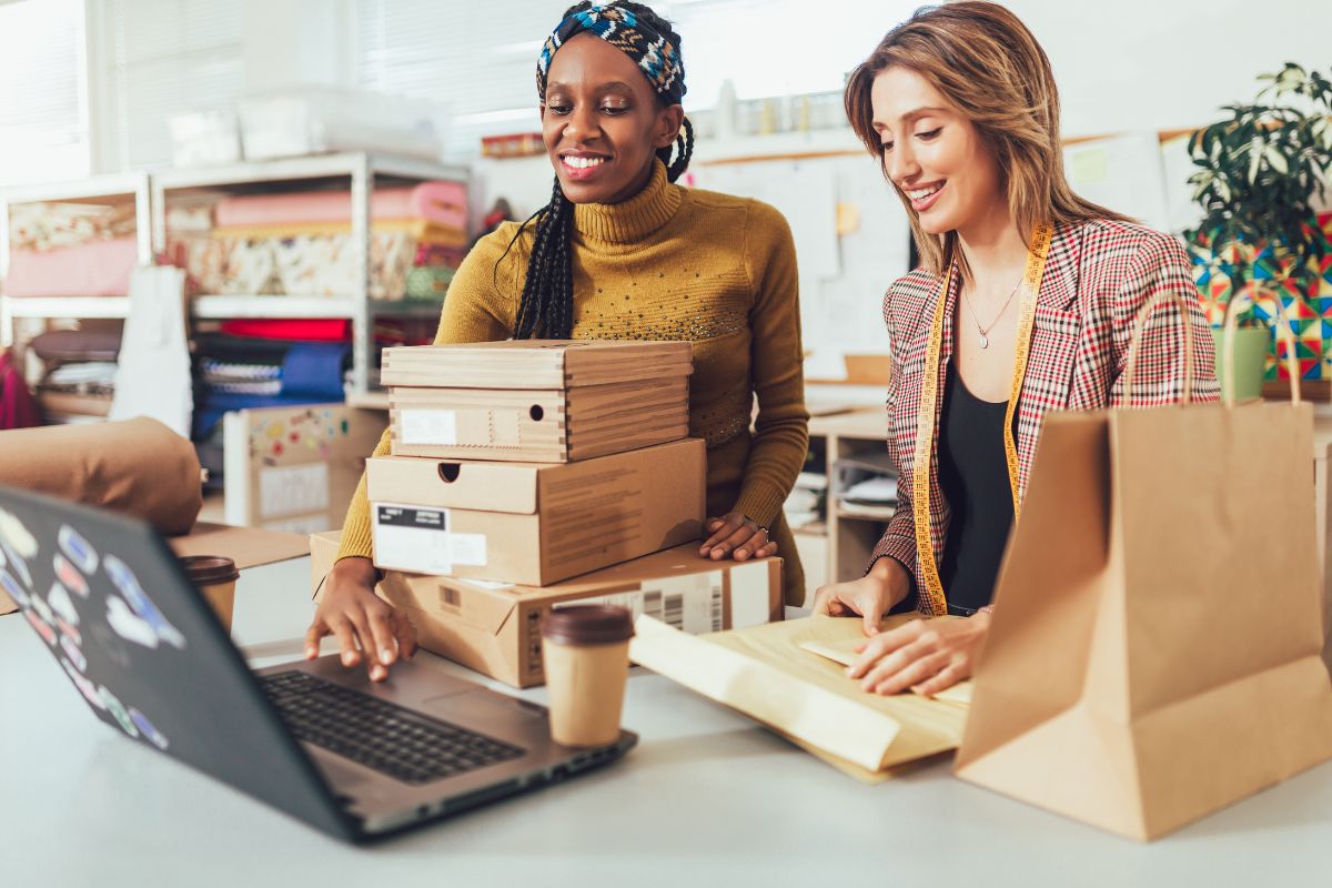 Two women listing products on Etsy shop using laptop along side cardboard boxes on the table