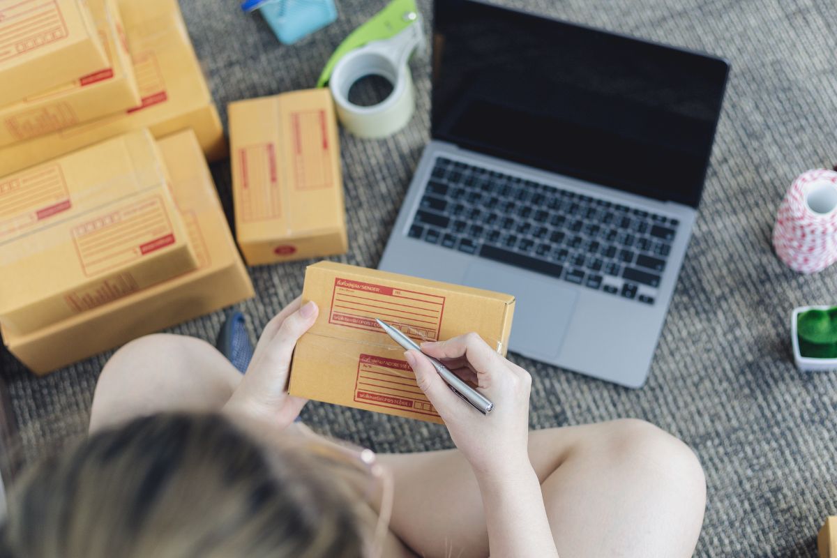 Woman writes on cardboard box surrounded by laptop and products for online selling