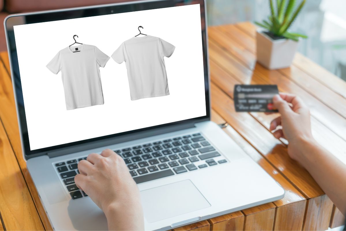 A man holds a credit card while using design software on laptop to craft t shirt