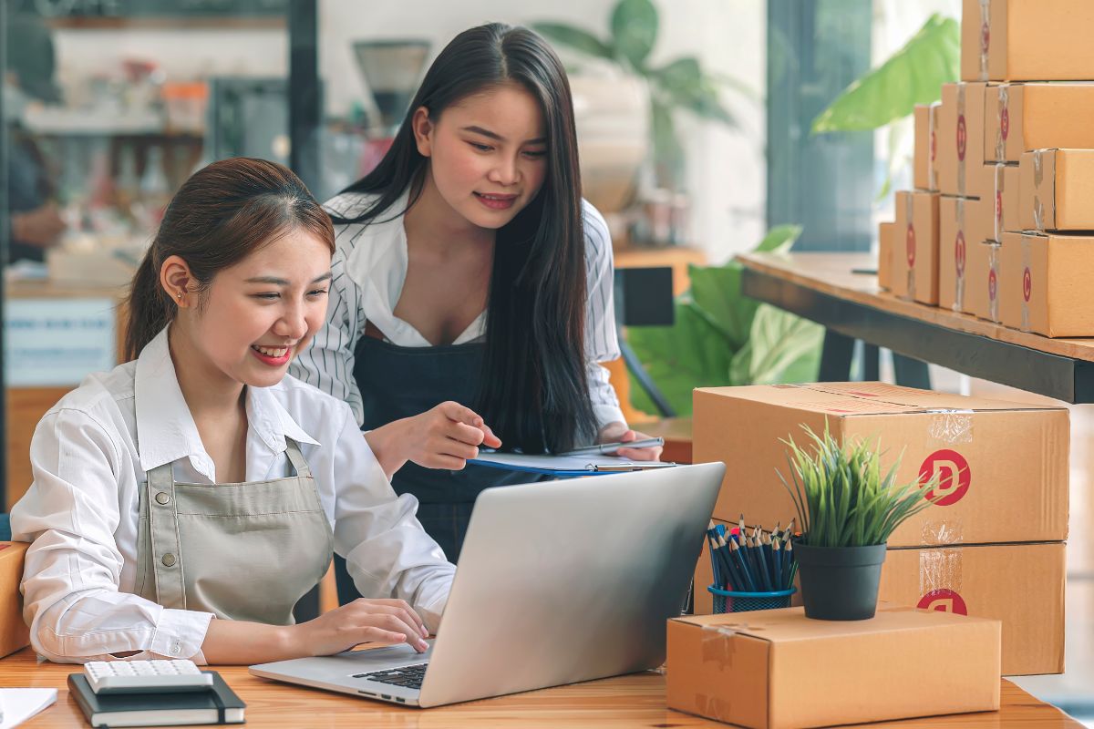 Two woman working on laptop alongside cardboard boxes on the table to market their print on demand business